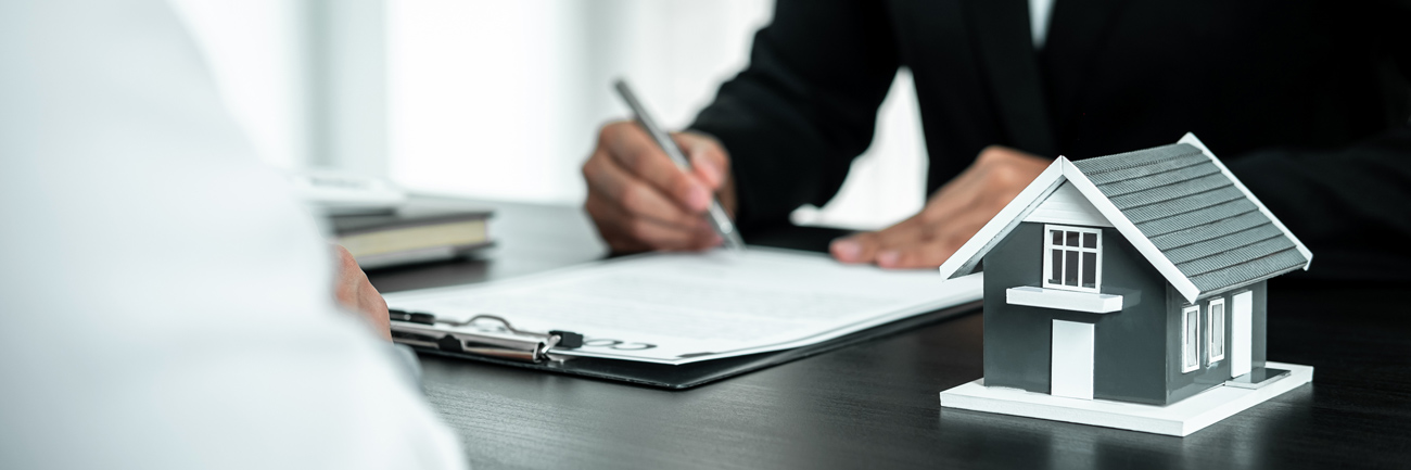 A person signing home loan insurance paper with house model and key on table